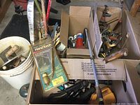 White plastic bucket filled with wooden-handled hand tools such as trowels and putty knives. Also visible cardboard box containing other tools and supplies.