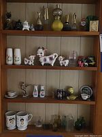 Overall shelf view showing ceramic cow figurines, vases with floral designs, mugs, animal figurines, and perfume atomizers arranged on three wood-paneled shelves.