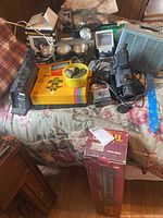 Photo showing the assortment of vintage cameras, a movie camera, projector, Polaroid camera, flash units, and VHS tapes on a floral bedspread.