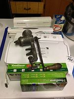 Overall view of the vintage food grinder alongside Foodsaver vacuum sealer, vacuum bags, and Mason jar caps on a white surface.