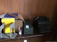 Black Royal manual typewriter, metal Cole card file cabinet, yellow metal file keepers, grey metal bookends, rulers, pencils in clear cup on brown wooden surface against wood panel wall