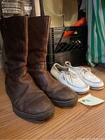 Side view of a pair of tall brown leather boots and a pair of white low-top leather shoes on the floor with a label marked 'A'.