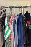 Clothing rack showing various women's tops and blouses plus a handbag, including a green and white striped short-sleeve blouse, coral pink polka dot blouse, floral blouse, light blue blouse, and dark navy blouse.