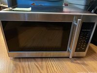Front view of the stainless steel Hamilton Beach microwave oven showing the door, handle, and digital control panel on a wood floor.