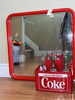 Red pipe mirror with curved edges reflecting floor and staircase, alongside red Coke bottle carrier with bottles inside