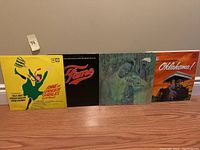 Four vinyl record albums laid out side by side on the floor. Covers visible: Anne of Green Gables, Fame, Nat King Cole, Oklahoma!.