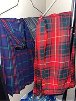 Three large plaid skirts/kilts on hangers showing distinct patterns and colors: blue with green and red, gray with white, and red with green and yellow fringed edge.