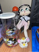 Photo showing a glass jar with Mickey and Minnie Mouse artwork, white lid, Betty Boop plush, Furby toy, and a small glass construction worker figure with orange helmet on a wooden surface.
