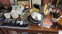 Wide view showing collection of white ceramic bowls and ramekins, packaging cutlery, metal mixing bowls, bundt pan, kitchen utensils in ceramic holder plus decorative kitchen plaques and signs.
