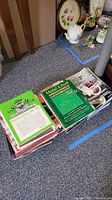 Two piles of assorted books on carpet near decorative porcelain items. Top book is Model A Ford Shop Manual Vol 1 in green cover.