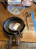 Three cast iron fry pans stacked, clear Pyrex mixing bowl, Pyrex measuring cup, and metal scissors on wooden table.