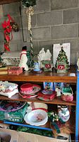 Photo showing Christmas serving dishes, tins, figurines including a snowman and various holiday-themed plates and tins arranged on shelves against a concrete block wall.