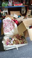 Photo showing Nutcracker figure, white Santa's workshop sack, box of assorted Christmas ornaments, cardboard boxes, and shelves with other boxed decorations and lights.