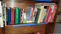 Cookbooks arranged on a wooden shelf featuring diverse titles with colorful covers, some spiral bound and others hardcover or paperback.