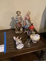 Four figurines displayed on a wooden surface including man with shovel, woman with basket, cardinal bird, and reclining child, along with a candle holder and bowl.