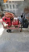 Side view of red Briggs & Stratton generator with orange and black extension cords coiled on top and attached to the generator frame, positioned against a block wall in a shed environment.
