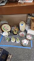 Overview of hummingbird-themed lot items arranged on floor including plates, figurines, vase, and teapot