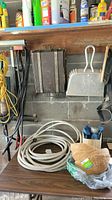 Photo showing coiled white garden hose, metal dust pan with brush, measuring cup, coco liners for plant pots, and black carrying case on a workbench with concrete block wall background.