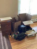 Wide shot showing the filing cabinet, Fellowes shredder, Canon printer, and various office supplies packed in a cardboard box and on the floor in a room with window and hardwood floor.