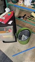 Overall view of black plastic storage drawers with paint rollers, brushes, scrapers on shelving in background. Green plastic bucket with paint roller inside on floor.