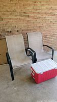 Two beige mesh patio chairs with black metal frames positioned side by side against a brick wall. A red and white Coleman cooler is in front.