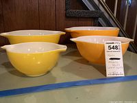 Four vintage Pyrex mixing bowls: two yellow and two orange with sunflower designs, arranged on a surface. Tags indicate largest bowl size 13x14 inches.