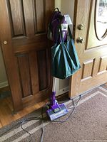 Photo of the full upright Shark Rocket vacuum cleaner in purple color leaning against a door, showing its entire height, vacuum head and cord.
