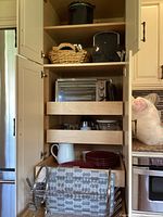 Kitchen cabinet with multiple small kitchen appliances including a black and stainless air fry toaster oven, a coffee maker on pull-out shelves, stacked red plates, white ceramic pitcher, and a metal dish drainer rack.