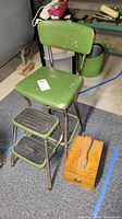 Green Cosco step stool chair with metal frame and vinyl seat and backrest, showing white stains and wear, next to wooden shoe shine kit box with attached brushes.