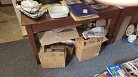 Wooden table with various household items placed on top. Boxes are stored underneath. Table surface and legs visible, showing reddish-brown finish.