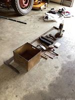 Wide shot showing all items laid out on concrete floor near garage door including wooden box, saw, lantern, truck manual, thermometer, wrenches, ruler, tractor headlight