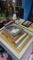 Photo showing a variety of picture frames stacked on a table, including a large gold decorative frame, silver and black frames, and some framed prints.
