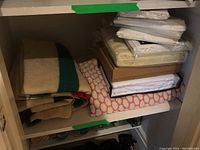 Shelf view showing two folded Hudson’s Bay point blankets left, red/white geometric textile bottom right, stacked white bedding sets top right and beige folded piece middle right