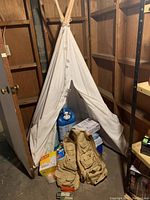 Photo showing small white canvas teepee tent supported by wooden poles, fishing vest and water jugs, boxes of trash bags and cups below tent in basement corner.