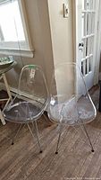 Two clear acrylic chairs with metal legs placed side by side on wooden floor near white wall and door.