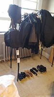 Metal clothing rack with black denim jackets, other dark clothing, and two black bags hanging. Several pairs of shoes arranged on the floor.