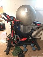 Full view of the Trainer 5008 stationary bike with resistance bands on the bike handlebars, large silver exercise ball on top, dumbbells and kettlebell on the floor, and weight plates nearby.