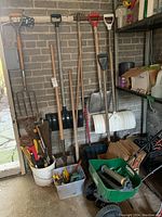 Full view of garden tools and accessories against brick wall including shovels, rakes, buckets with smaller tools, turf builder spreader and cardboard sheets