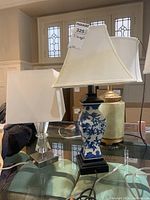 Three tabletop lamps: two with blue floral ceramic bases on dark wooden square pedestals and fabric shades, one clear acrylic and metal base lamp with a square white fabric shade, displayed on glass table.