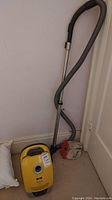 Full view of yellow canister vacuum with attached hose and wand leaning against wall