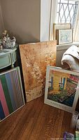 Three framed prints leaning against a wall and furniture: a tall floral branch print, a colorful striped print, and a garden scene print.