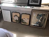 Four framed prints shown side by side on floor: an abstract print with silver frame, two water lily floral prints in black rounded-edge frames, and a cubist style portrait in a brown wooden frame.