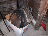 Box containing multiple silver-plated platters and trays stacked vertically showing various shapes and decorative edging, placed in basement corner.