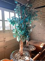 Two tall faux leafy plants with braided wood-like trunks in ornate dark metal urn-style containers filled with decorative stones, shown near a window and partially inside a garage space.