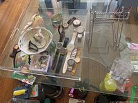 Photo showing wrist watches, ceramic bowl with jewelry, acrylic tray, and metal plate stands on a glass table