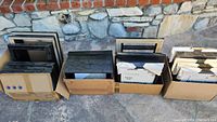 Wooden picture frames stacked inside three cardboard boxes placed outdoors near stone wall. Frames are mostly black with some natural wood tones. Sizes vary but many near 11" x 14".