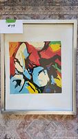 Framed colorful abstract painting by Eric Fiazi, 17 x 21 inches, shown with visible white mat and silver frame on patterned rug background.