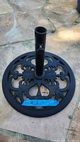 Photo shows a circular black metal umbrella stand base with ornate floral cutout patterns and central cylindrical tube for umbrella placement.
