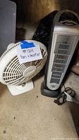 Photo showing a white tabletop fan and a gray tower stand heater placed on a concrete floor in a storage setting.
