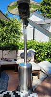 Full view of a stainless steel outdoor heat lamp set on a patio with cushioned outdoor furniture and greenery in the background.